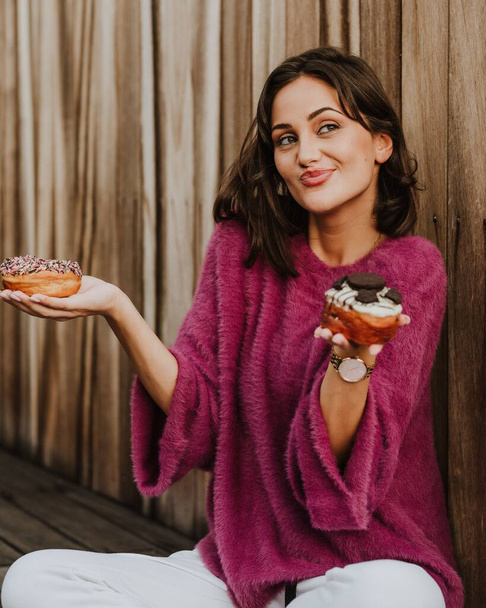A smiling girl holding two delicious donuts, contemplating which to eat first - Foto, imagen