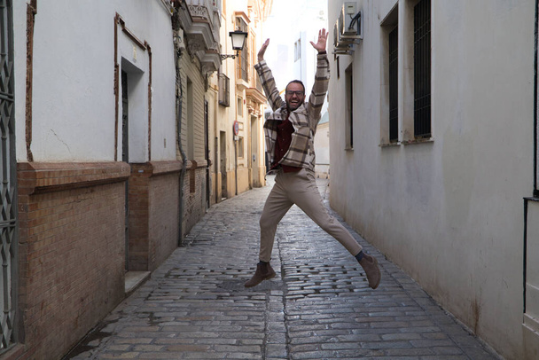 A handsome young man with a beard and glasses is jumping for joy in a narrow street. Happiness and joy concept. - Foto, immagini