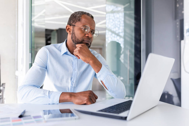 Thinking serious African American businessman working inside office, successful business man boss looking at laptop screen pondering important technical decision. - Foto, Imagen