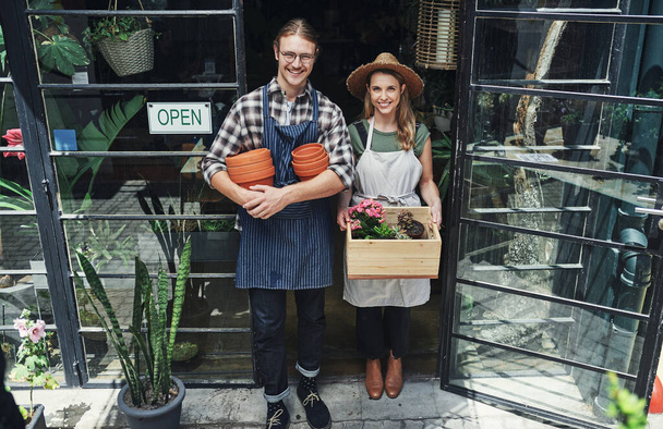 Rośliny są w doniczce. Portret dwóch młodych przedsiębiorców stojących razem przy wejściu do ich kwiaciarni i trzymających artykuły do doniczkowania - Zdjęcie, obraz