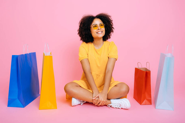 ショッピングコンセプト。幸せな素敵なアフリカ系アメリカ人やブラジル人の若い女性の完全な長さの写真,黄色の夏のドレスで,隔離されたピンクの背景に紙袋の間に座って,離れて見て,笑顔 - 写真・画像