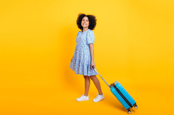 Volledige lengte van een gelukkig mooi Afrikaans Amerikaans of Braziliaans meisje met krullend haar, in een blauwe zomerjurk, een reiziger, staat op een geïsoleerde gele achtergrond met een blauwe koffer, kijkt weg, glimlacht - Foto, afbeelding