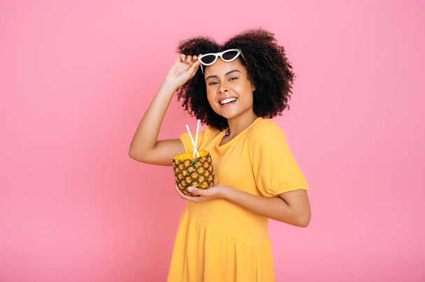 Lovely african american or brazilian curly woman, in yellow summer dress, holds a pineapple in hands, fresh pineapple tropical cocktail, smiling at the camera, stand on isolated pink background - Photo, image