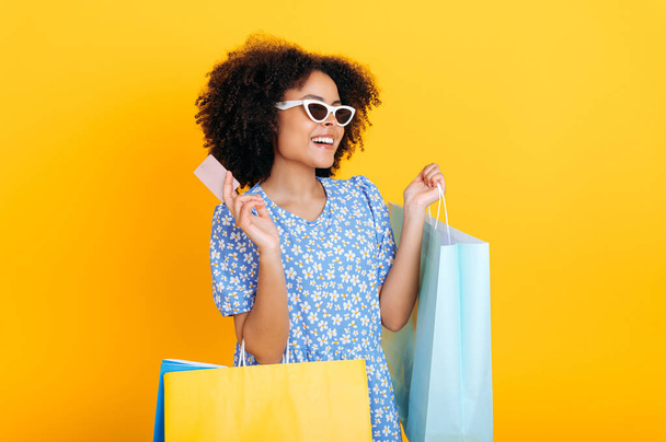 Excited happy lovely curly haired african american woman in an orange summer dress, with glasses, holds shopping bags and credit banking card, looks away, smiles, stand on isolated orange background - Fotó, kép