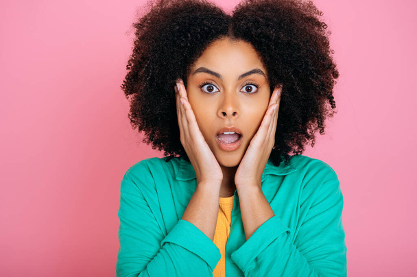 Amazed shocked african american or brazilian curly haired beautiful young woman in green shirt, looking at camera in surprise, holding palms near cheeks and opening her mouth, isolated pink background - Photo, image