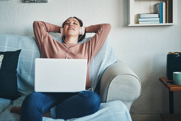 Este día es para hacer lo que quiera. una mujer joven usando un ordenador portátil y auriculares mientras se relaja en el sofá en casa - Foto, imagen