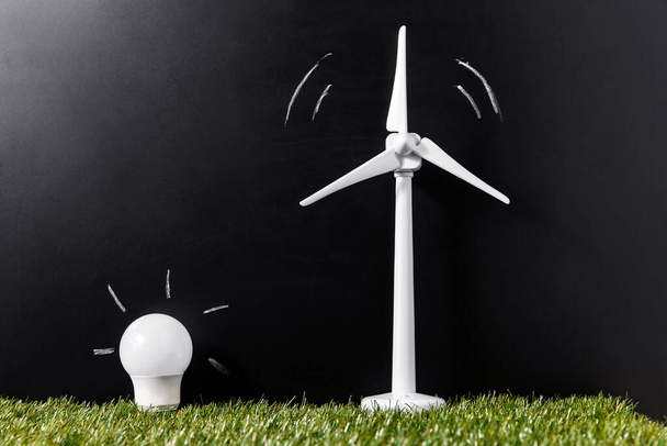 power, ecology and sustainability concept - close up of wind turbine model and energy saving lightbulb on green grass over black background - Foto, immagini
