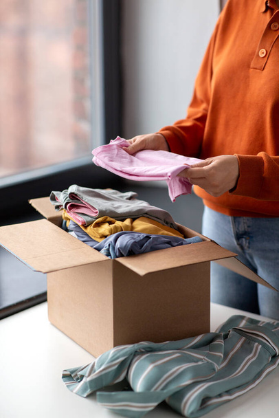 charity, donation and volunteering concept - young woman puts clothes in cardboard box at home - Foto, immagini