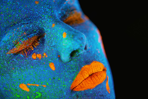Neon verf, make-up en vrouw gezicht close-up met donkere achtergrond en creatieve cosmetica. Surrealistische gloed, fantasie en psychedelische cosmetica van een vrouwelijk model met unieke en creatieve kunst in de studio. - Foto, afbeelding