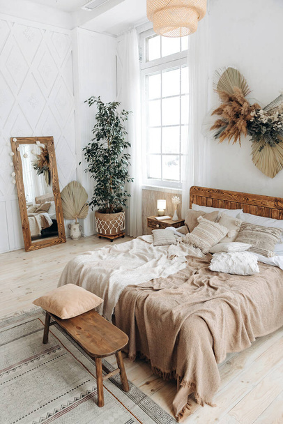 Cozy rustic bedroom with boho ethnic decor. Bright spacious apartment with large windows. Wooden furniture. Large mirror. Handmade textile. Plants in the interior. Nobody. - Foto, Bild