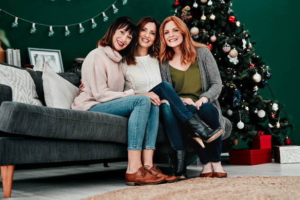 Beste Freunde für immer. Porträt von drei attraktiven Frauen mittleren Alters, die zusammen auf einem Sofa sitzen und zu Hause einen Weihnachtsbaum im Hintergrund haben - Foto, Bild