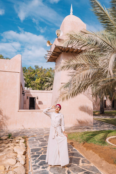 Feliz chica asiática en pañuelo para la cabeza caminando en el casco antiguo o pueblo patrimonio en los Emiratos Árabes Unidos - Foto, imagen