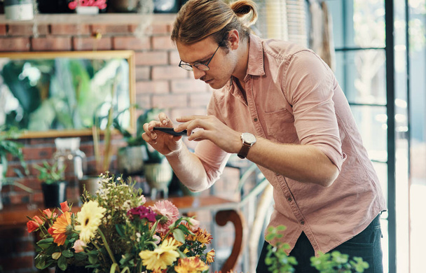 Estos se verán muy bien en mis redes sociales. un joven hombre de negocios guapo parado en su floristería y usando su celular para fotografiar sus flores - Foto, Imagen
