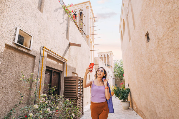 With her smartphone in hand, a happy girl tourist snaps a selfie to share with her followers from the charming old town of Dubai. - Photo, Image