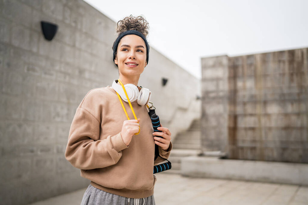 Portrait of one young adult caucasian woman stand outdoor in front of modern concrete wall with headphones confident with rubber resistance band tubes copy space - Zdjęcie, obraz