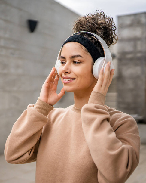 Kafasında kulaklıklarla modern beton duvarın önünde duran genç bir beyaz kadının portresi müziğe güveniyor. - Fotoğraf, Görsel