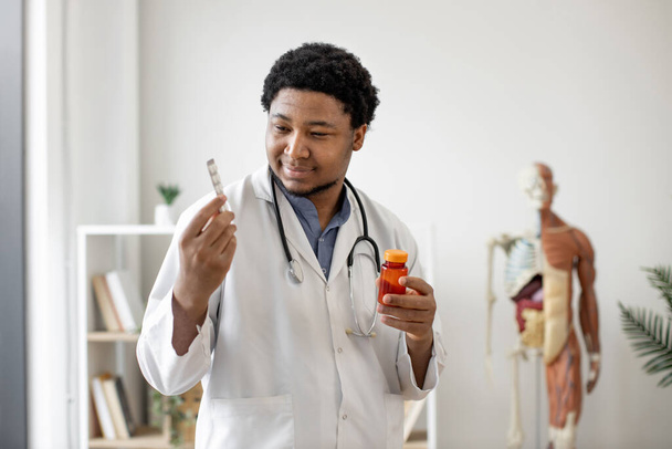 Friendly multiethnic man in lab coat scanning pill bottle while standing with tablets in doctors workplace against anatomical model background. Efficient pharmacist studying prescription drugs. - Fotoğraf, Görsel