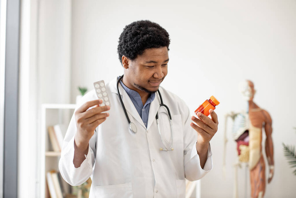 Friendly multiethnic man in lab coat scanning pill bottle while standing with tablets in doctors workplace against anatomical model background. Efficient pharmacist studying prescription drugs. - Фото, зображення