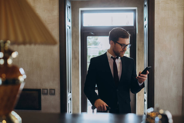 Businessman walking in hotel lobby with suitcase and using his smart phone. Male business traveler in hotel hallway with cellphone and luggage. - Zdjęcie, obraz
