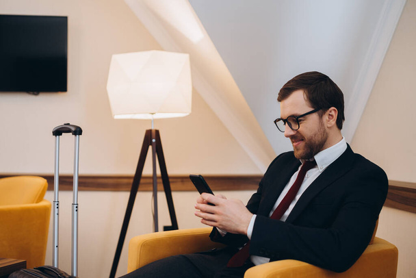Egy üzletember ellenőrzi a telefonját, miközben egy kényelmes székben ül egy hotelszobában.. - Fotó, kép
