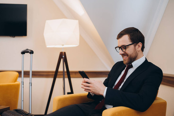 Um homem de negócios verifica seu telefone enquanto se senta em uma cadeira confortável em um quarto de hotel. - Foto, Imagem