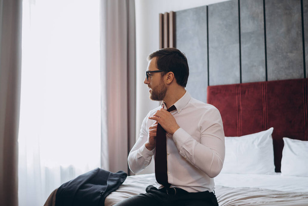 Mladý podnikatel si sundává kravatu, zatímco sedí na posteli v hotelovém pokoji. - Fotografie, Obrázek