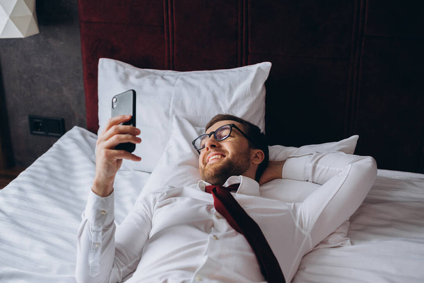 Ein Geschäftsmann liegt auf einem Bett in einem Hotelzimmer. Ein junger Mann benutzt sein Smartphone während er ruht. - Foto, Bild