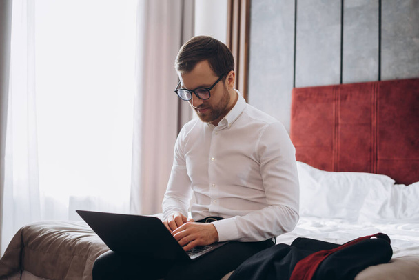 コンピュータを扱う若いビジネスマンがホテルの部屋のベッドに座っている - 写真・画像