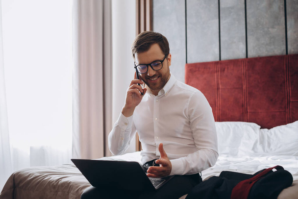 Όμορφος επιχειρηματίας που κάνει phonecall ενώ κάθεται στο κρεβάτι δωμάτιο του ξενοδοχείου και χρησιμοποιώντας φορητό υπολογιστή κατά τη διάρκεια επαγγελματικό ταξίδι - Φωτογραφία, εικόνα