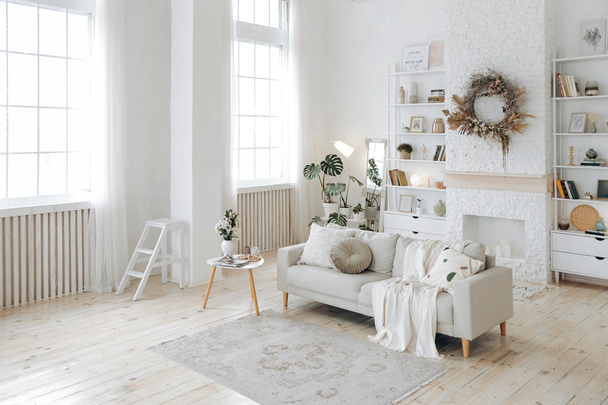 Moderna y elegante sala de estar blanca con grandes ventanales y sofá de estilo escandinavo con el telón de fondo de una chimenea y estanterías y plantas en maceta. Nadie.. - Foto, Imagen