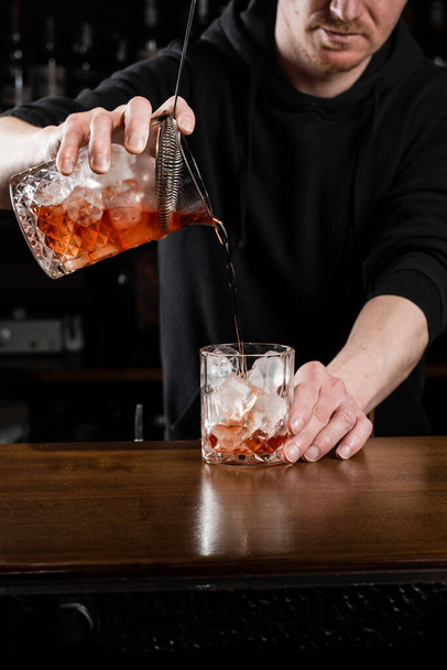 Bartender pours mixed traditional Negroni cocktail in the glass at the bar. Bartender mixes gin, campari, and sweet vermouth to prepare the Negroni alcoholic cocktail - Фото, изображение