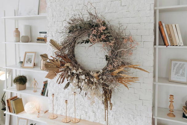 A wreath of dried flowers over the fireplace between shelving with books and decor in a stylish modern Scandinavian living room. Nobody. - Photo, Image