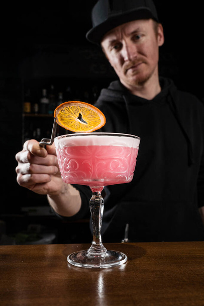 Μπάρμαν διακοσμεί ροζ Clover Club αλκοολούχο κοκτέιλ με πορτοκαλί φέτα στο μπαρ. Μπάρμαν αναμιγνύει ασπράδι αυγού, λεμόνι, ξηρό βερμούτ και τζιν για να προετοιμάσει το Clover Club κοκτέιλ - Φωτογραφία, εικόνα