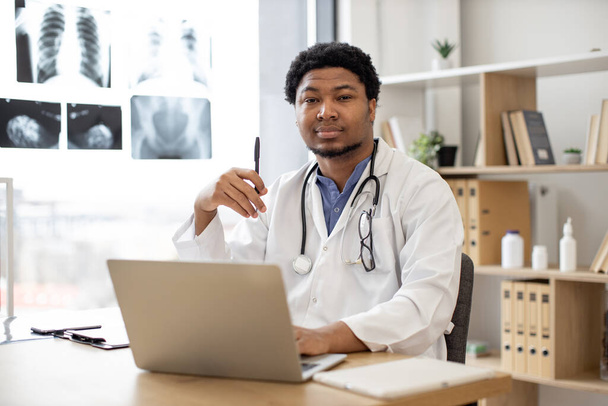 Pensive multiethnic male therapist in white coat posing with pen in hand behind computer on writing desk in doctors office interior. Healthcare practitioner exploring diseases using technologies. - Foto, Imagem