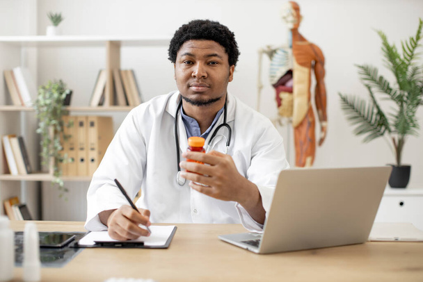 Mindful multiethnic man wearing stethoscope around neck writing on clipboard while examining medicine bottle on anatomical model background. Therapist taking notes on pills using computer in office. - Фото, зображення