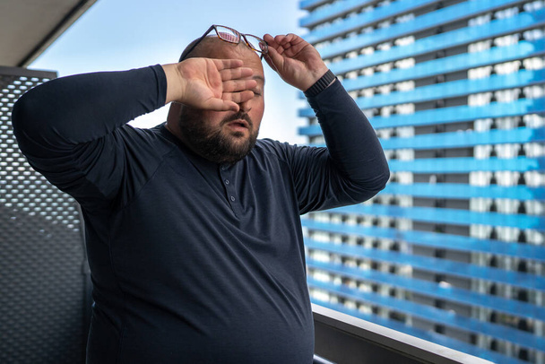 Trpí nadváhou muž cítí bolest hlavy tření očí stojící na balkóně za horkého počasí v létě. Vyčerpaný samec snáší tlakové výkyvy žijící v tropickém podnebí s vysokou vlhkostí. Zdravotní problémy. - Fotografie, Obrázek