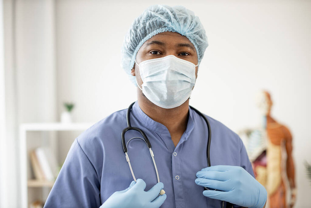 Portrait of african american male in blue scrubs wearing face mask, medical cap and latex gloves holding stethoscope around neck in workplace. Health professional getting ready for examination. - Фото, изображение