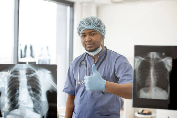 Friendly multiracial male in scrub top and latex gloves giving thumb-up gesture while staying behind clear wall panel with X-ray scans. Happy surgeon showing approval after successful operation. - Photo, image