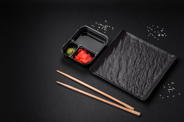Pickled ginger, soy sauce and wasabi in portioned plastic containers on a dark concrete background - Фото, изображение