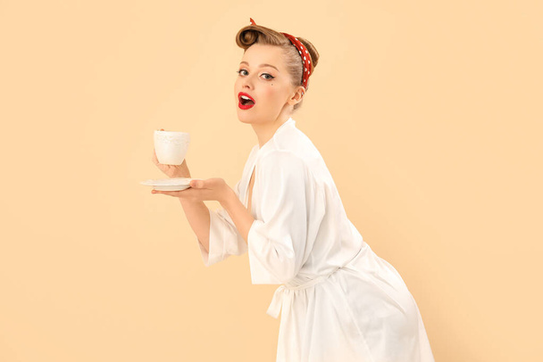 Young pin-up woman with cup of coffee on beige background - 写真・画像