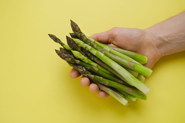 Fresh green asparagus in hand on yellow background, top view. copy space. Healthy food for balanced diet. - Фото, изображение
