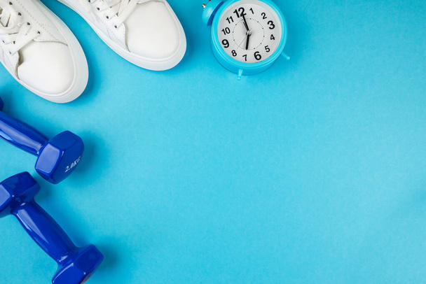 Fitness accessories concept. Top view photo of white alarm clock sneakers and blue dumbbells on isolated blue background with copy space. It's time to train. - Valokuva, kuva