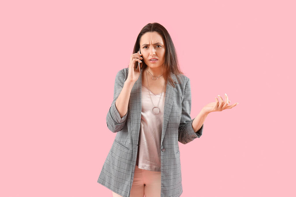 Displeased young woman talking by phone on pink background - Fotó, kép