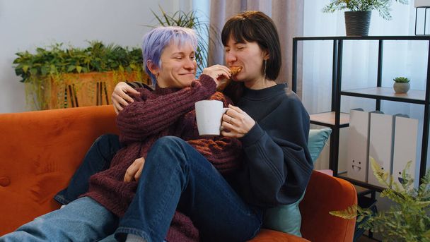 Two lesbian women family couple, girl friends drinking coffee or tea, eating croissants and talking at home morning living room. LGBT same-sex people. Enjoying happy breakfast together lying on sofa - Photo, image