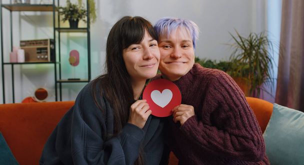 Due giovani donne lesbiche coppia di famiglie o ragazze amiche abbracciano tenendo forma di cuore dalla carta, segno dei social media, mostrando amore a casa. Donna omosessuale che si abbraccia. Parità delle persone LGBT. gay, bisessuale - Foto, immagini