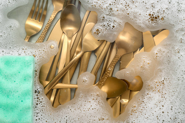 Cleaning sponge with golden cutlery and foam in water, closeup - Photo, Image