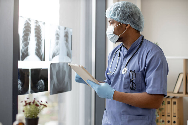 Hombre multiétnico atento en matorrales médicos y máscara facial mirando imágenes de rayos X mientras sostiene la tableta en el lugar de trabajo de los médicos. Médico en guantes y gorra examinando escáneres torácicos en el interior del hospital. - Foto, Imagen