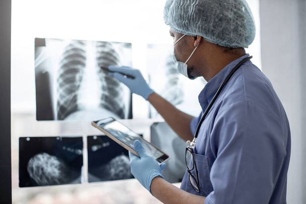 Fókuszáljon a mellkas röntgen vizsgálatára, amelyet multiraciális egészségügyi szakember tanulmányozott védőöltözetben, injekciós tollal ellátott tablettát használva. Férfi radiológus azonosítására tömeg a bal tüdőben technológiát alkalmazó. - Fotó, kép