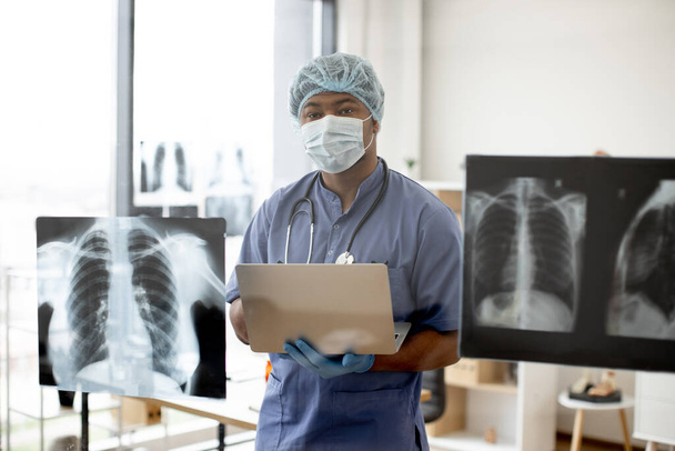 Focused multiethnic male in medical scrubs, cap and mask holding computer while standing in front of glass panel with chest scans on it. Experienced physician diagnosing medical disease in hospital. - Photo, image