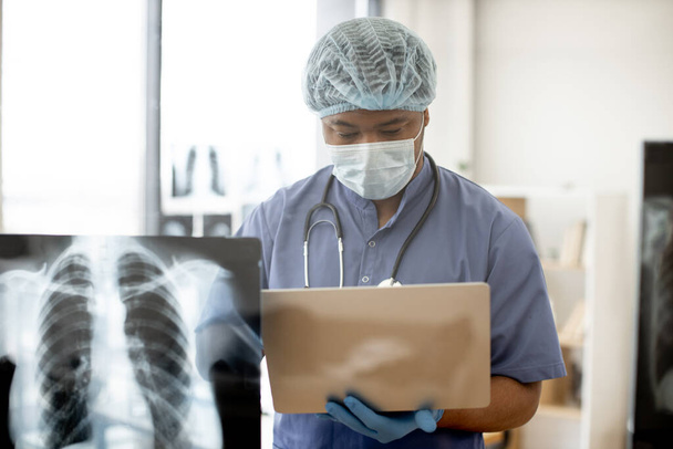 Focused multiethnic male in medical scrubs, cap and mask holding computer while standing in front of glass panel with chest scans on it. Experienced physician diagnosing medical disease in hospital. - 写真・画像